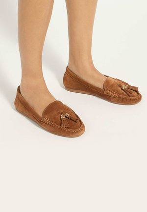 Mocassins marron en daim à enfiler avec des détails à pompons portés aux pieds sur un fond neutre.