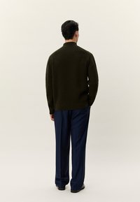 Ember sötétzöld, texturált pulóvert és tengerészkék nadrágot viselve, háttal áll egy sima, világos háttér előtt.