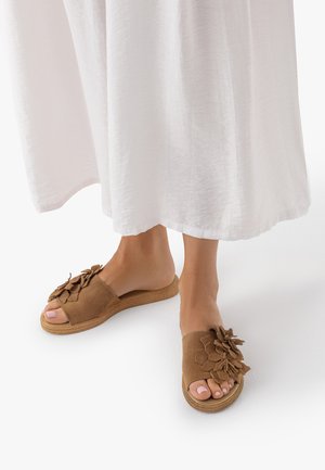 Stopy w brązowych sandałach wsuwanych z ozdobami kwiatowymi, w połączeniu z długą białą spódnicą, stojące na białym tle.