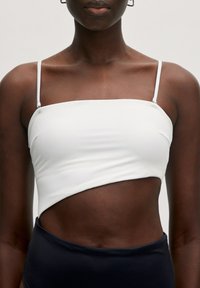 Witte crop top met smalle bandjes, voorzien van een diagonale snit en een gladde textuur, gecombineerd met donkerblauwe high-waisted onderkleding.