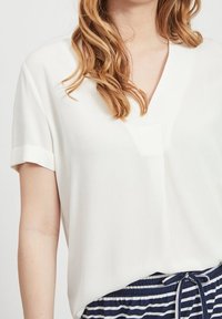 Blouse blanche à manches courtes avec un col en V, présentant une texture lisse. Associée à un pantalon rayé bleu marine à la taille.
