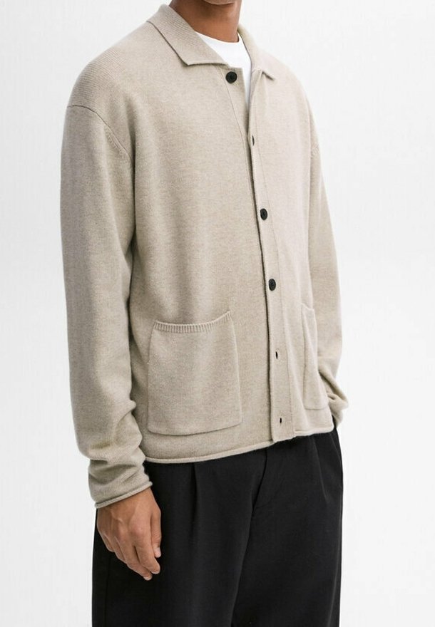 Beige gebreide cardigan met een kraag, een sluiting met knopen aan de voorkant, twee grote zakken en geribbelde manchetten. Draagt over een wit shirt.