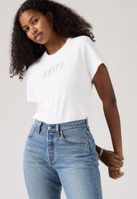 T-shirt blanc à manches courtes avec le logo "LEVI'S" encadré de métal, associé à un jean taille haute bleu clair avec passants de ceinture et poches avant.