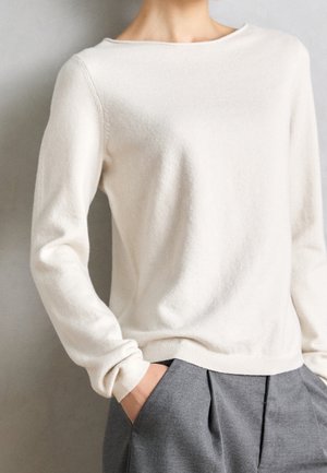 Donna che indossa un maglione lungo a maniche lunghe color crema, leggermente infilato in pantaloni grigi plissettati, con una mano in tasca, su uno sfondo semplice.