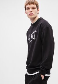 Μαύρα φούτερ φτιαγμένο από μείγμα βαμβακιού, με λευκή γραφική εκτύπωση στο στήθος, ριχτό λαιμό και μανίκια. Χαλαρή εφαρμογή με στρωτό σχέδιο.