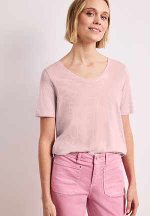 Blonde Frau trägt ein hellrosa kurzärmeliges Shirt mit Rundhalsausschnitt, das in hohe pinke Jeans gesteckt ist und steht vor einfachem Hintergrund.