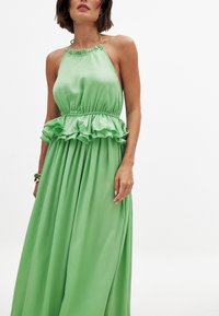 Vihreä satiininen halterneck-mekko, jossa on röyhelöpeplum-yksityiskohta, laskostettu hame ja säädettävä kaulahihna. Liukas pinta, sulava muotoilu.