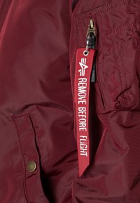 Bordeaux nylon bomberjack met een ritszak en een rode tag met de tekst "VERWIJDER VOORDAT JE VLIGT" bevestigd aan de rits.