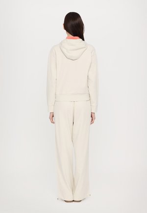 Hoodie color beige chiaro e pantaloni a gamba larga coordinati, entrambi realizzati in tessuto morbido. Il hoodie presenta un cordino e una foderatura interna vivace.
