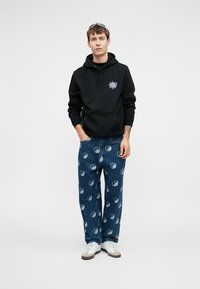 Fekete kapucnis pulóver színes grafikával, kék farmernadrággal, amelyen yin-yang minta található, és fehér sneaker cipővel.