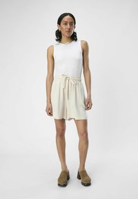 Hvid ærmeløs top parret med lysebeige trækbåndsshorts, der har et struktureret materiale, lårhøjde og løs pasform. Brune klipklapper fuldender looket.