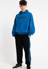 Niebieska bluza z kapturem z napisem "FEARLESS" w kolorze czarnym, w połączeniu z czarnymi spodniami dresowymi z niebieskimi paskami po bokach. Materiał wydaje się miękki i casualowy.