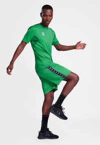Jersey atlético verde y pantalones cortos con acentos laterales negros, combinados con zapatillas y calcetines negros, mostrados en una pose en acción.