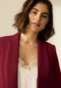 Sacou bordo cu rever îngust purtat peste un top crem cu dantelă la margine. Textură fină a țesăturii; croială ajustată cu linii clare.