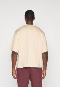 Béžové tričko s krátkym rukávom a okrúhlym výstrihom, vyrobené z hladkej tkaniny. Zadná časť má voľný strih, v kombinácii s vínovočervenými nohavicami.