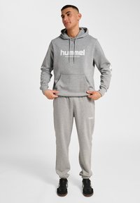 Szara bluza z kapturem i dresy wykonane z miękkiego materiału, z dużym białym logo i subtelnymi wzorami na rękawach, połączone z czarnymi butami.