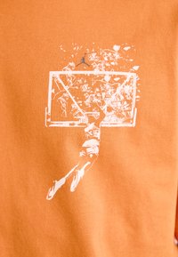 Orange stof T-shirt med en hvid grafik af en basketballspiller, der laver en dunk, og en kurv, med en lille blå Jordan-logo ovenfor.