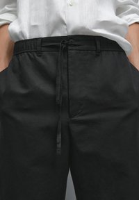 White buttoned shirt tucked into black pants with an elastic waistband and drawstring tied at the front. Hands in pockets.