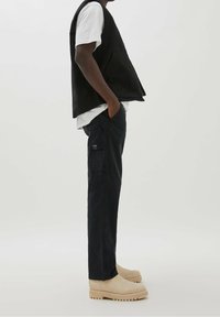 Chaleco negro sin mangas sobre una camiseta blanca, acompañado de pantalones cargo oscuros y botas beige. Incluye bolsillos laterales y un corte relajado.