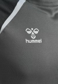 Tissu de sport texturé gris avec un logo d'abeille Hummel blanc et le nom de la marque près d'une couture avec des sections de couleur noire et blanche.