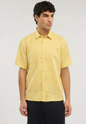 Hombre con camisa amarilla de manga corta abotonada y pantalones oscuros, de pie contra un fondo claro y liso, mirando hacia un lado.