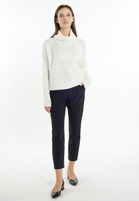 Witte, ribbelige trui met een hoge kraag, gecombineerd met slim-fit zwarte broek en glanzende zwarte hakken. Model staat tegen een effen achtergrond.