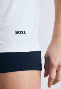 Balta kokvilnas T-krekls ar tumši zilām apmalēm, kurā ir melna izšuvuma “BOSS” logo tuvu apakšmalai. Tuvplāns uz rokas un apakšējās apģērba malas.