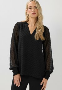 Donna bionda che indossa una blusa nera a fantasia con maniche a sbuffo trasparenti e polsini arricciati, che posano contro un muro chiaro e semplice.