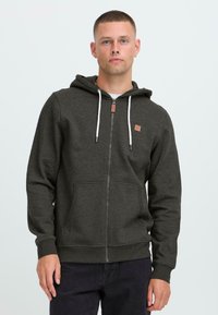 Man with short hair wearing dark grey zip-up hoodie with white drawstrings and black pants standing against a plain light background.