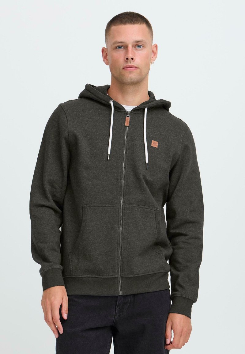 Man with short hair wearing dark grey zip-up hoodie with white drawstrings and black pants standing against a plain light background.