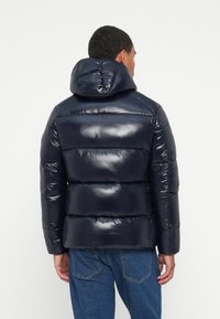 Man wearing a shiny navy blue puffer jacket with a hood, paired with blue jeans, facing away against a plain white background.