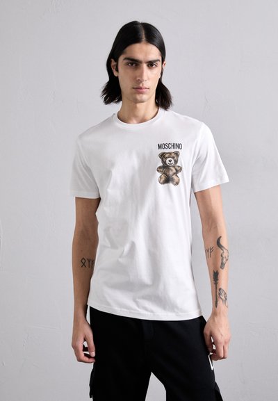 Hvid bomulds t-shirt med et print af en bjørn og "Moschino" tekst på forsiden, korte ærmer, rund halsudskæring, båret med sorte bukser.