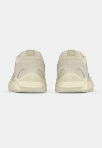 Pár béžových športových topánok zobrazených zozadu, s hrubými podrážkami a závesnými pútkami na pätných častiach proti jednoduchému pozadiu.