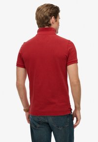 Polo rouge avec col à revers, manches courtes, et fentes latérales à l'ourlet. Le tissu semble texturé et respirant.