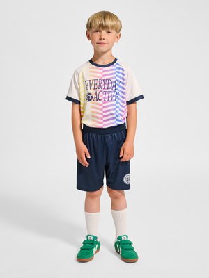 Młody blond chłopiec stoi nieruchomo, ubrany w wielokolorową koszulkę "EVERYDAY ACTIVE", granatowe spodenki, białe skarpety do kolan oraz zielone trampki.