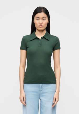 Σκούρο πράσινο πόλο με ριμπ ύφασμα, γιακά, κοντά μανίκια και λοξό κούμπωμα με τρία κουμπιά. Φορεμένο με ανοιχτό μπλε ψηλόμεσο τζιν.