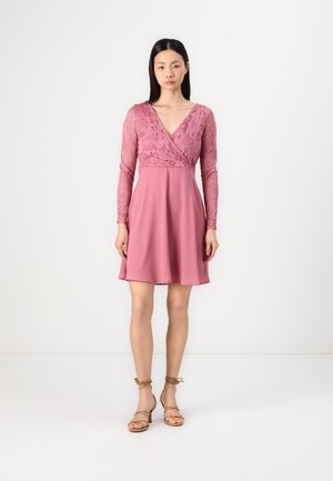 Robe rose pâle avec de longues manches en dentelle, un décolleté en V et une jupe évasée. Associée à des sandales nude à lanières, se tenant sur un fond uni.