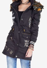Giacca parka nera con cappuccio foderato in pelliccia, toppe decorative, tasche con zip e polsini a coste. Presenta dettagli metallici e scritte.