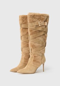 Botine înalte din suede bej, cu vârfuri ascuțite, tocuri stiletto, căptușeală pufoasă și curele încrucișate fixate cu catarame aurii.