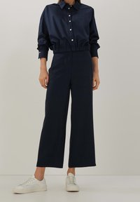 Marinblå skjorta med krage och elastisk nederkant samt knappstängning, ihop med vida marinblå byxor; stylat med vita sneakers.