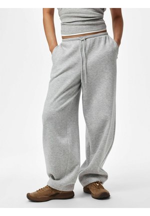 Personne portant un pantalon de survêtement gris ample, mains dans les poches et chaussures de randonnée marron, debout sur un fond blanc.