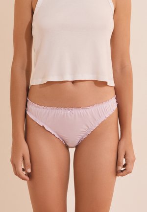 Femme portant un top court blanc et un bas de bikini rose clair à volants, debout sur un fond beige.