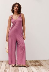 Kvinde med krøllet hår iført en ærmeløs, lyserød buksedragt og sandaler, holder en netpose med frugt og står på et trægulv.