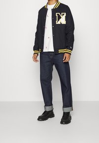 Svart varsityjacka med gula detaljer och en stor "N"-logotyp, vit T-shirt med små mönster, blå denimjeans och svarta stövlar.