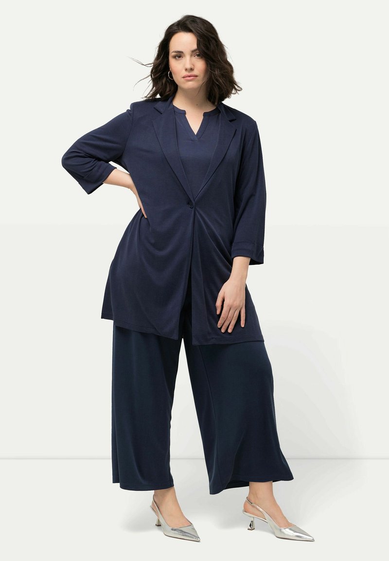 Marinblå, vida byxor i kombination med en matchande lång kavaj. Mjuk, fallande tyg med en enda knappstängning och ärmar som går till armbågen.