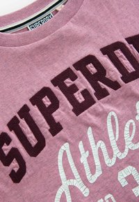 Rosa tyg T-shirt med mörk maroon "SUPERDRY" text och vit slitna "Athletics" bokstäver, med rund halsringning och mjuk konsistens.