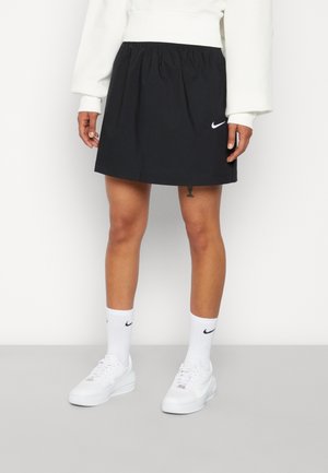 Personne portant une jupe noire Nike, un sweat blanc, des chaussettes blanches Nike et des baskets blanches, debout devant un fond uni.