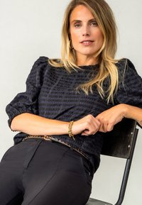 Zwarte textuur top met puffed mouwen, gedragen met zwarte broeken. Sieraden omvatten een gouden armband en een riem met decoratieve accenten.
