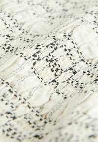 Gros plan sur un tissu blanc avec des motifs de coutures noires texturées créant un dessin ondulé et en relief.