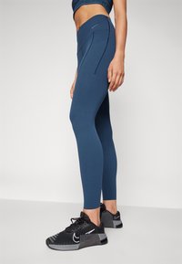 Navy blå leggings med høy midje, med kontrasterende sømdetaljer. Bært med svarte treningssko som viser et teksturert mønster.